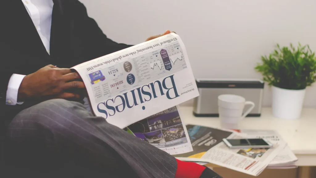 Person in a suit reading the business section of a newspaper beside a desk with coffee