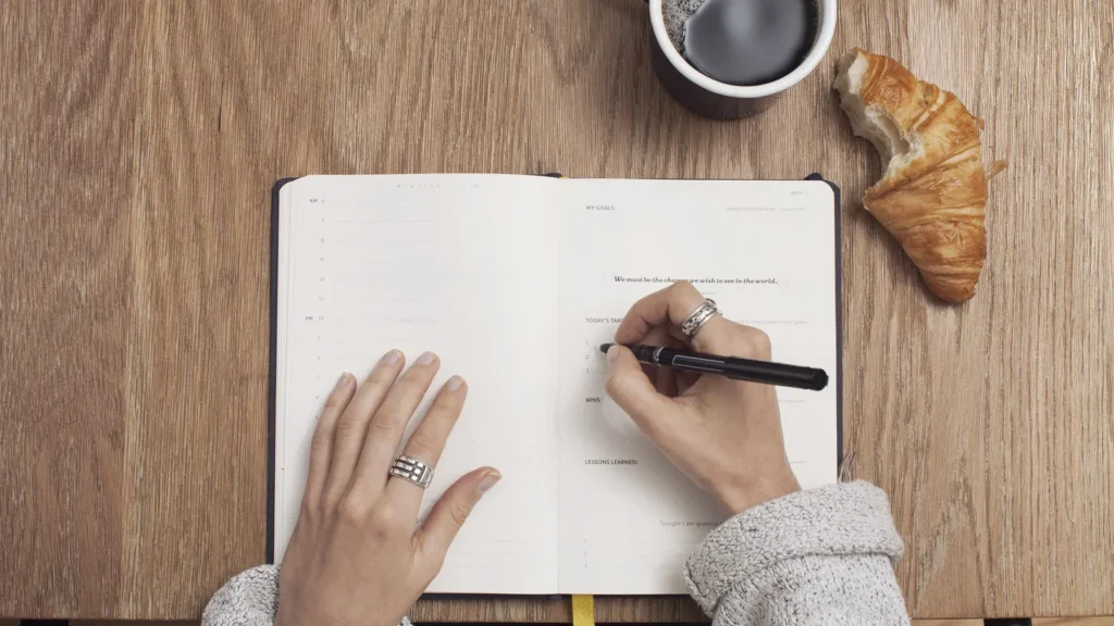 Hands writing in an open planner beside a coffee cup and croissant, noting keyword ideas