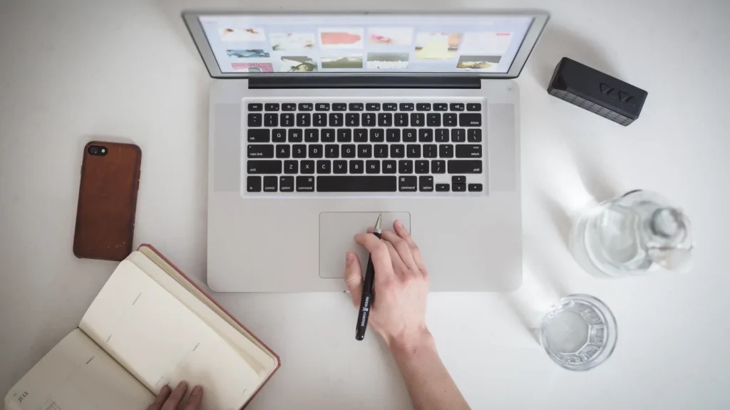 Top-down view of a laptop showing a website, with a hand holding a pen and an open planner beside it
