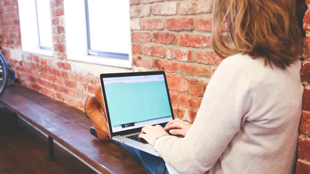 Woman working on a laptop by a bright window in a brick-walled workspace
